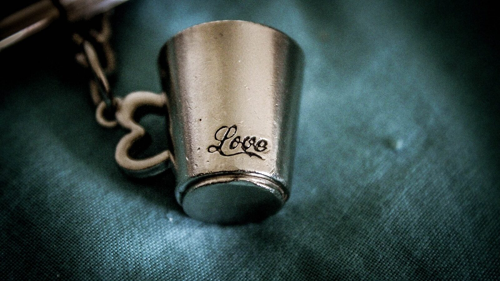 Silver keychain with a small cup design, inscribed with the word "Love," resting on a textured teal fabric background.