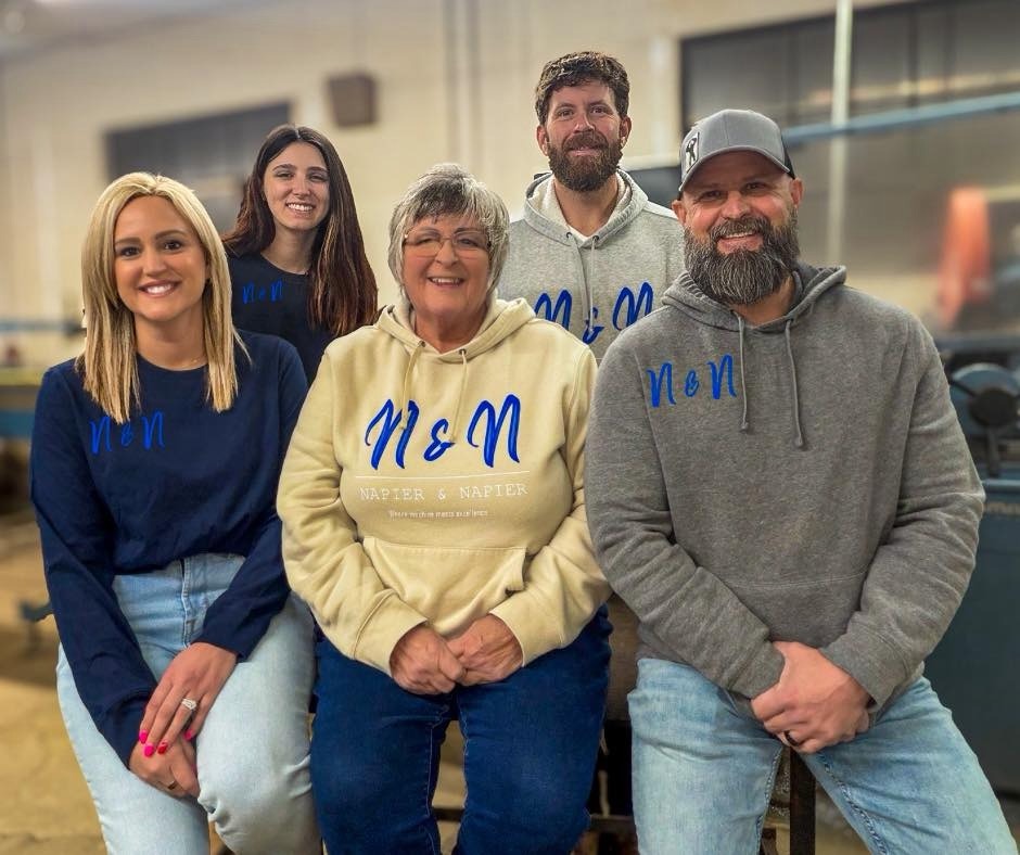 Five family members pose together in matching sweatshirts, smiling warmly. The backdrop hints at a workshop, showcasing a sense of togetherness and teamwork.